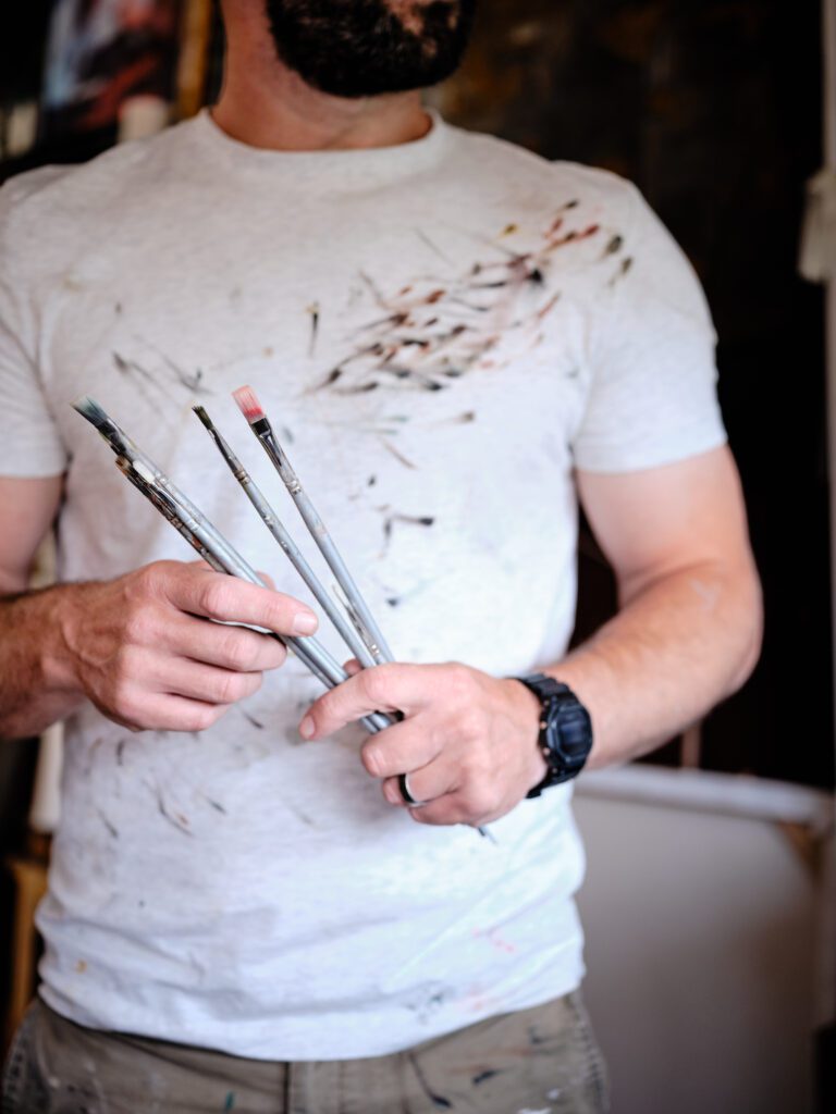 Will Rochfort is wearing a paint-stained white t-shirt and a black wristwatch is holding several Pro Arte Paintbrushes. The background is blurry, suggesting an art studio or workspace. Only the person's torso and hands are visible, reminiscent of a scene one might find in Will Rochfort's creative environment.A person wearing a paint-stained white t-shirt and a black wristwatch is holding several Pro Arte Paintbrushes. The background is blurry, suggesting an art studio or workspace. Only the person's torso and hands are visible.