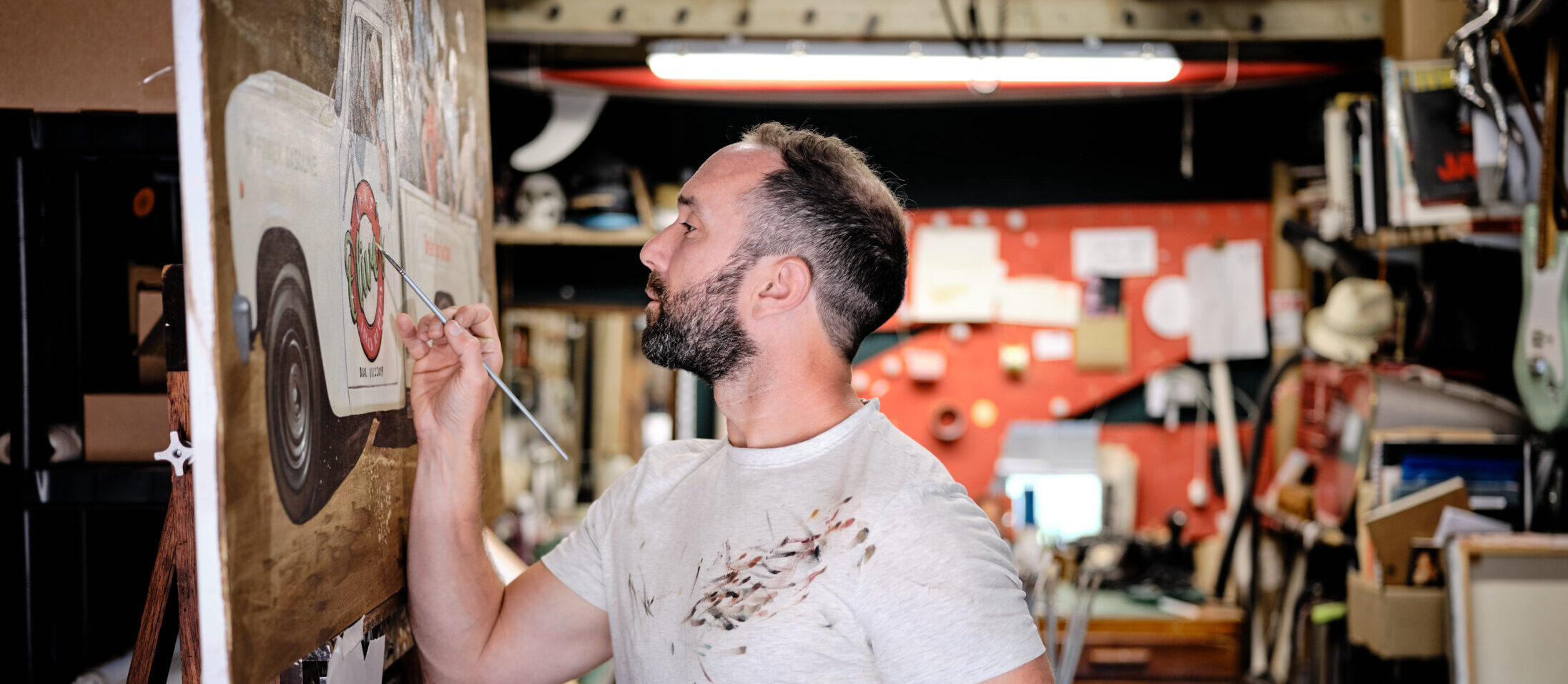 Will Rochfort stands in a cluttered workshop, painting a large canvas on an easel. He is wearing a light-colored, paint-splattered t-shirt and holding multiple Pro Arte Paintbrushes. The background features various art supplies, tools, and a bulletin board with notes and pictures of Will Rochfort’s works.