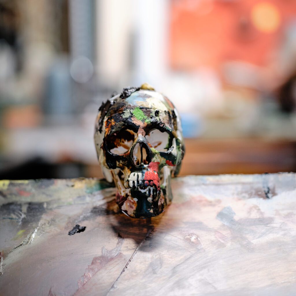 A small, colorfully painted skull sits on an artist's workbench. Paint brushes and blurry, abstract artwork can be seen in the background. The skull features a variety of bright colours and artistic patterns, suggesting a creative and vibrant workspace.