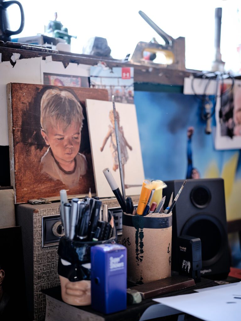 A cluttered artist's workspace features a painting of a young boy, brushes, and art supplies in containers, including Pro Arte Paintbrushes. Various tools are scattered about. Behind the setup, a vibrant blue and yellow painting by Will Rochfort hangs on the wall.