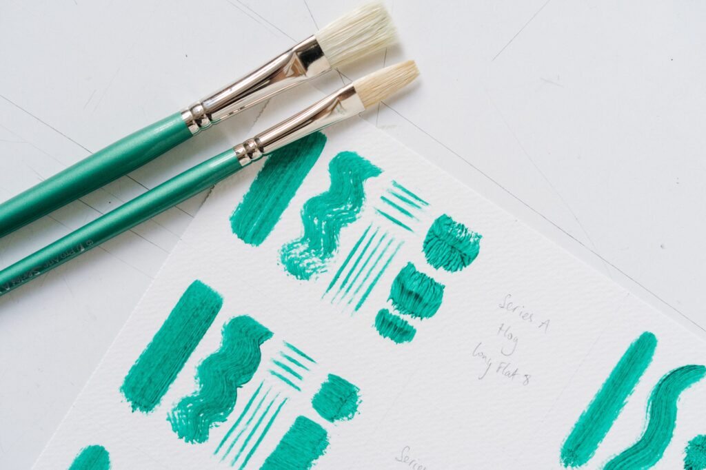 Two different types of paintbrushes next to various brushstrokes in green paint on white paper.