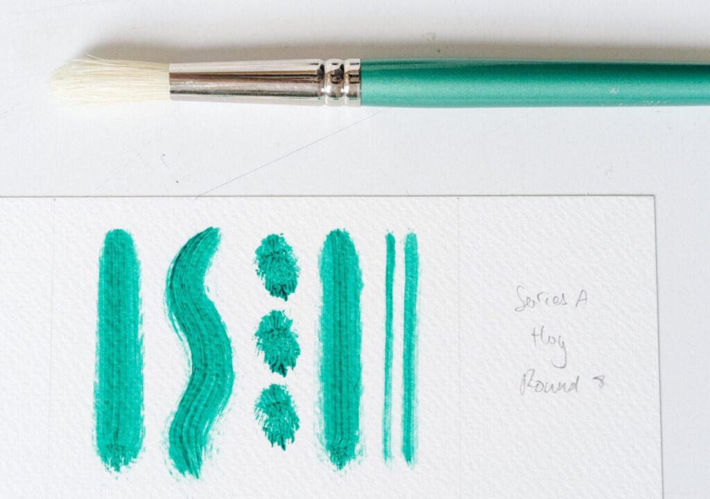 A single paintbrushes next to different brushstrokes in green paint on white paper.