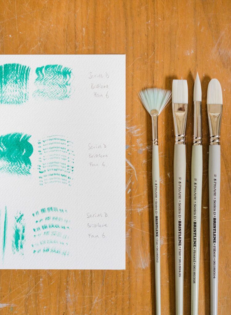 Four Pro Arte Brushes of varying shapes are laid out in a row on a wooden surface next to a piece of paper with green paint swatches and brush strokes. The paint swatches demonstrate different textures and patterns, labeled with the brush series and type.