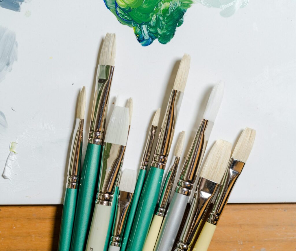 Different types of paintbrushes all different sizes, overlapping a piece of paper with some abstract painting in green and a wooden surface.