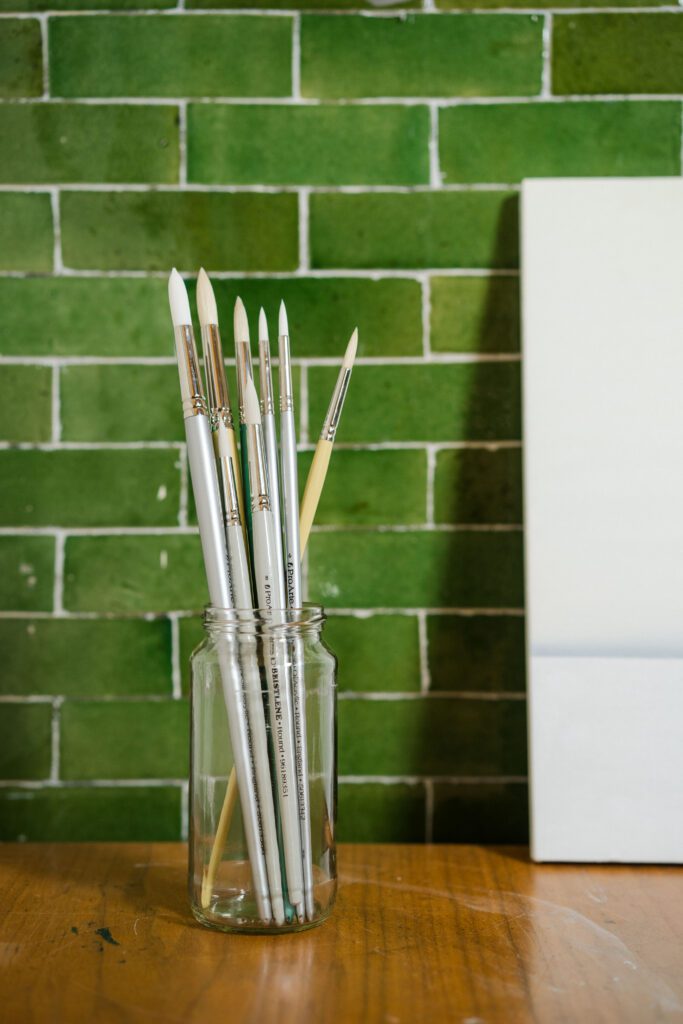Different types of paintbrushes in a glass jar, against a green tiled background.