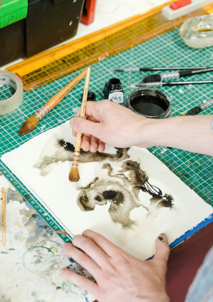 Images shows a close up of someones hands painting abstract patterns in green using a paintbrush
