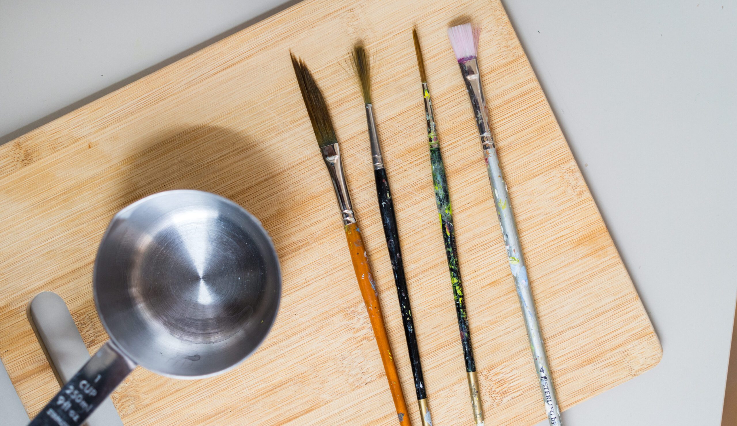 Image shows multiple paintbrushes of different shapes and sizes side by side on a wooden surface, alongside them is a small metal pot of water