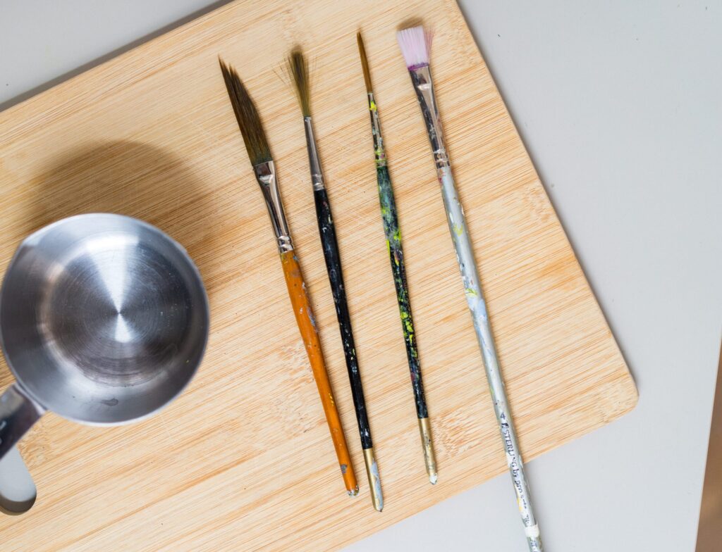 Image shows multiple paintbrushes of different shapes and sizes side by side on a wooden surface, alongside them is a small metal pot of water