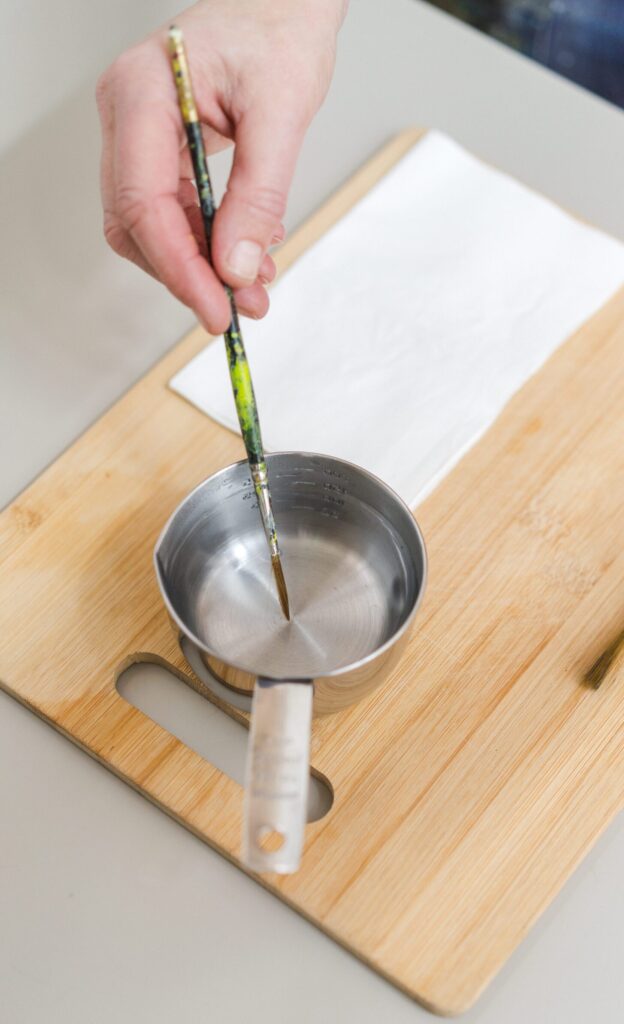 Images shows up a close up of an artist with a blank sheet of paper and a paintbrush in a metal container of clear water