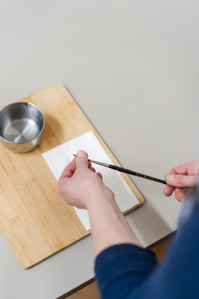 Images shows up a close up of an artist with a blank sheet of paper, a metal container of clear water and the artist is reshaping the brush bristles with their fingers