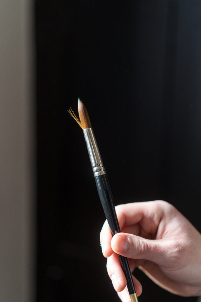 A close-up of a hand holding a paintbrush with a black handle and a metal ferrule. The brush bristles are slightly fan-out and illuminated by light against a dark background.
