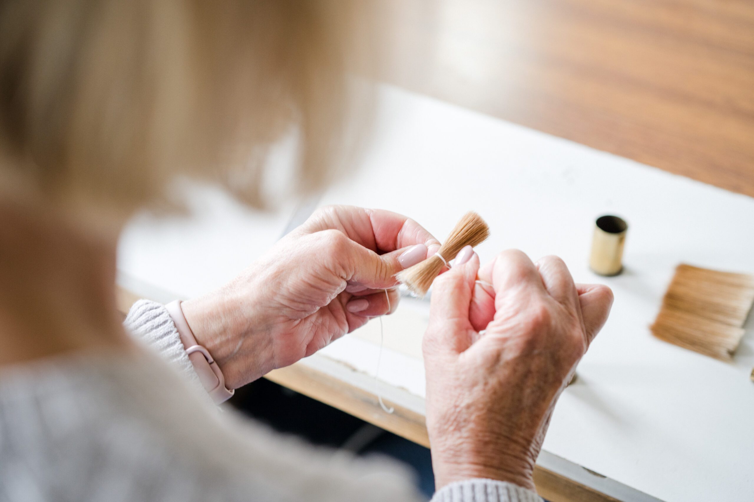 A person with light hair is shown from behind, tying a small bundle of sable bristles with a white thread. The individual is sitting at a table with a spool of thread and more bristles visible in the background.