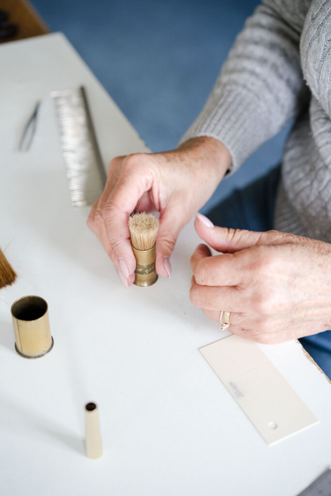 A person wearing a grey sweater is holding the bristles which will be used to make a Pro Arte paintbrush. On the table, there are a card, an open container, and a metal comb. The person's hands are in focus, revealing that they are involved in a task.