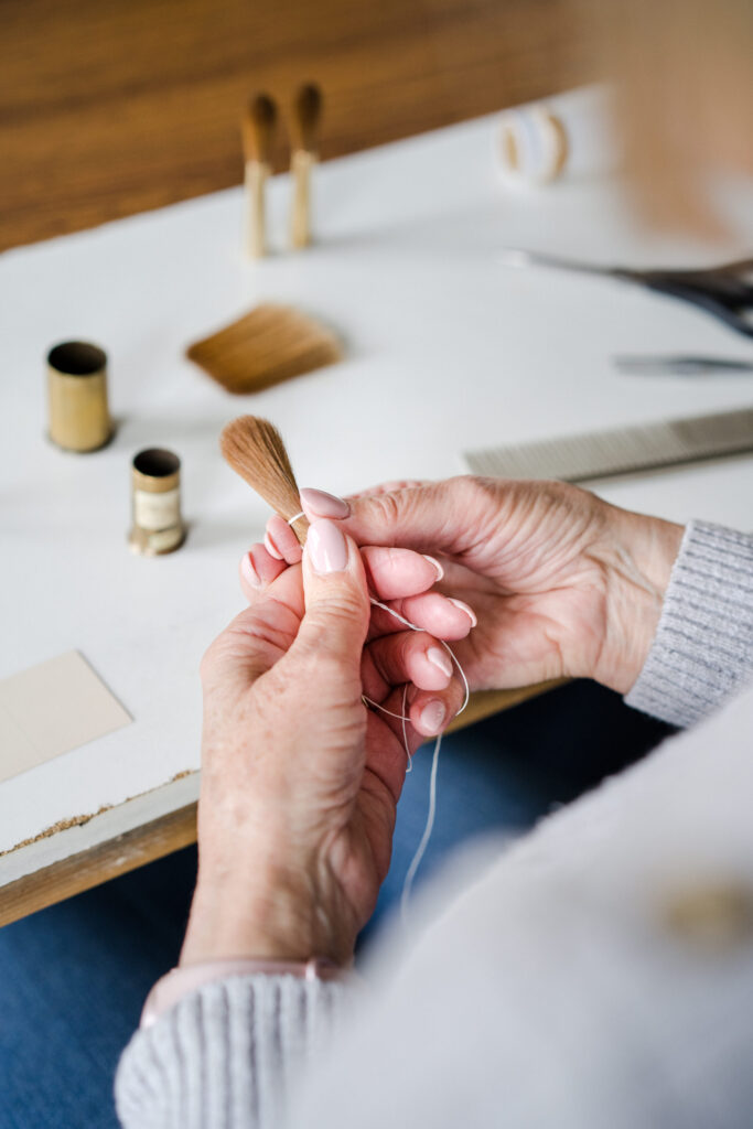 A person with light hair is shown from behind, tying a small bundle of sable bristles with a white thread. The individual is sitting at a table with a spool of thread and more bristles visible in the background.