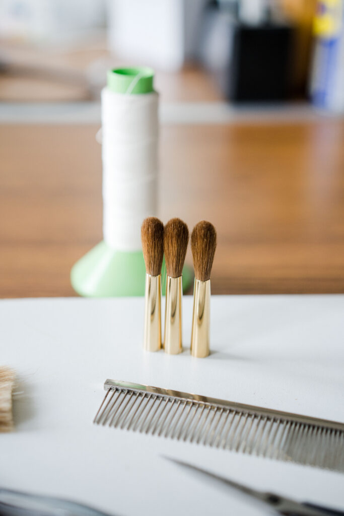 A close-up of three pro arte brush heads with light brown bristles standing upright in a row. The brushes have gold-colored ferrules. In the background, there is a spool of white thread, and in the foreground, there is a metal comb, scissors and paintbrush hairs.