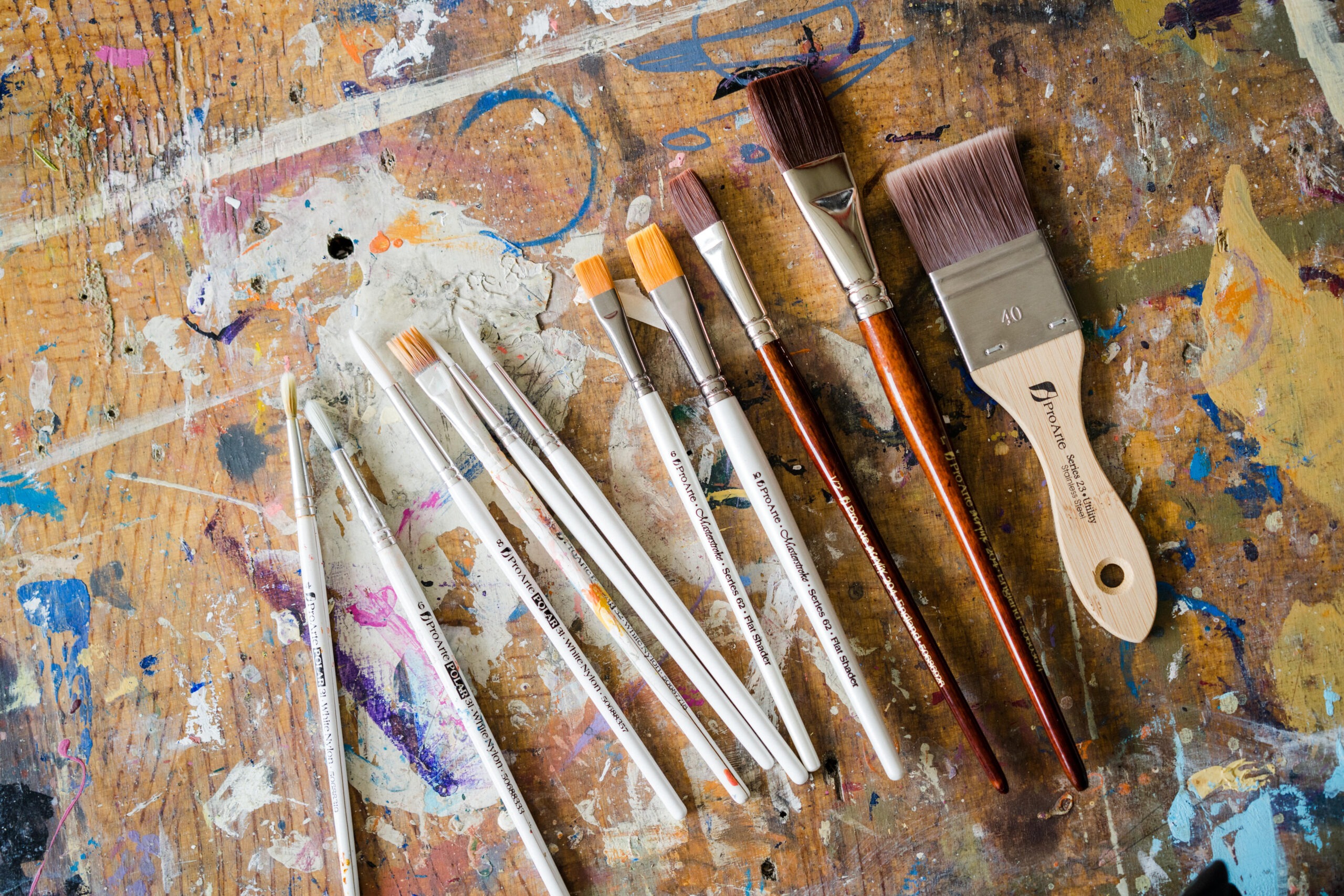 Pro Arte Paintbrushes of different sizes and shapes are laid out on a paint-splattered wooden surface. The brushes range from thin to wide bristles, indicating varied uses in painting. The colourful and messy background suggests a creative workspace.