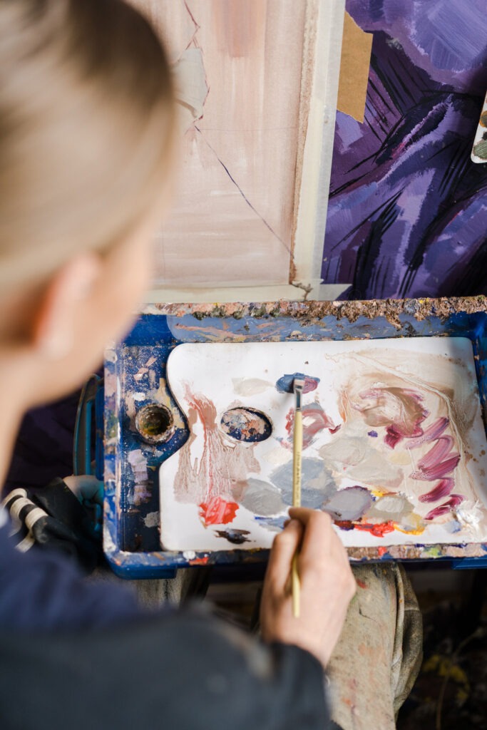 A young artist with long blonde hair uses Pro-Arte brushes while working on an oil painting in a creative studio environment.