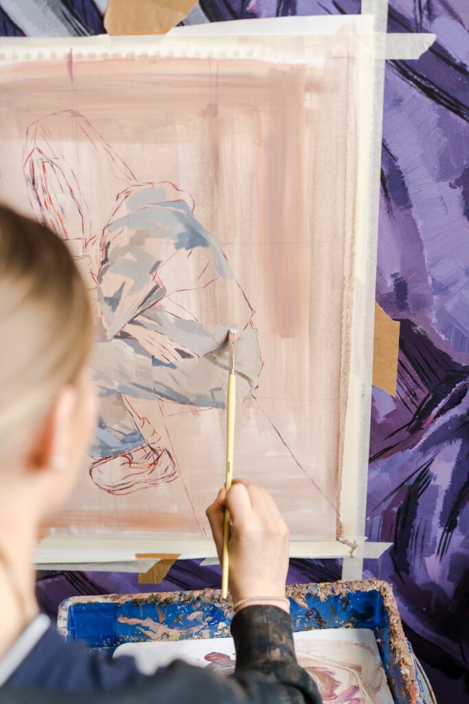 A young artist with long blonde hair uses Pro-Arte brushes while working on an oil painting in a creative studio environment.