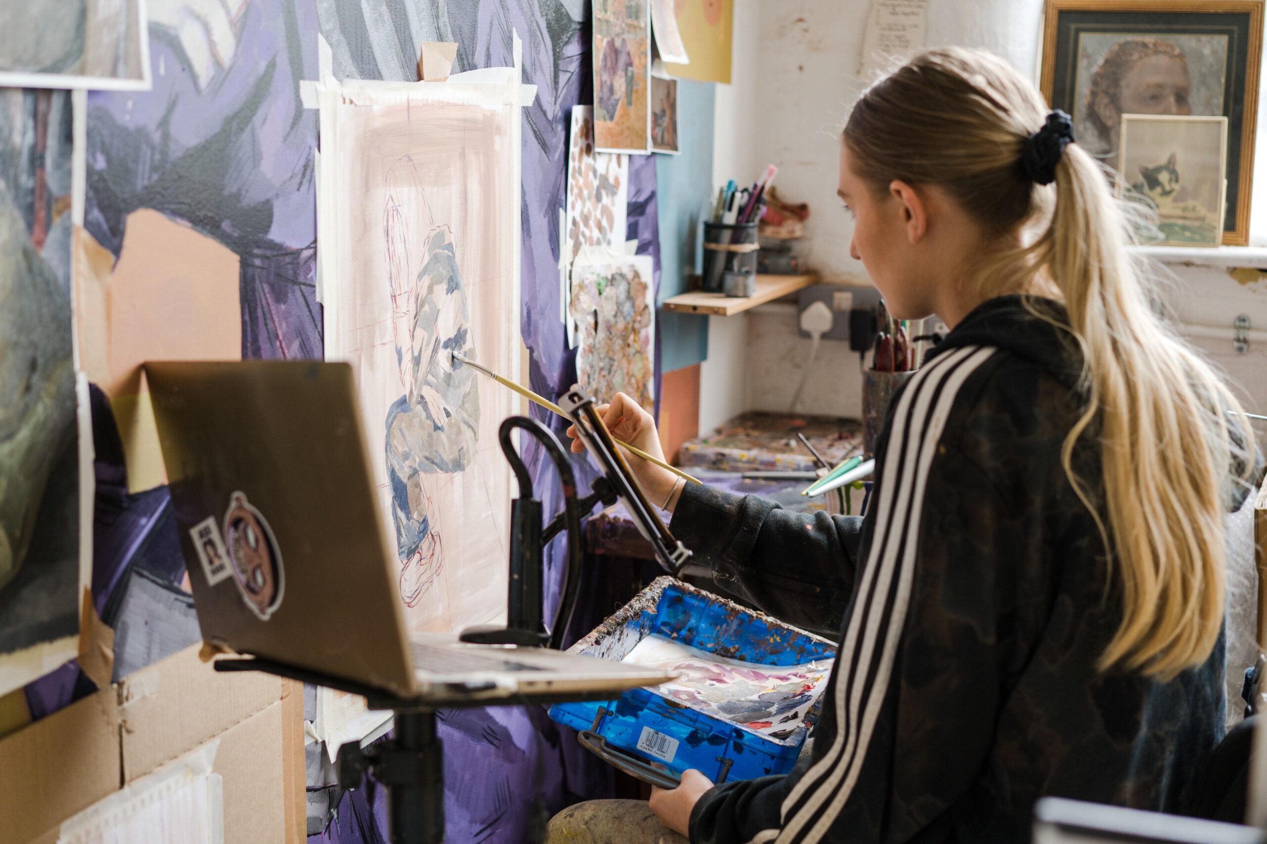 A young artist with long blonde hair uses Pro-Arte brushes while working on an oil painting in a creative studio environment. A laptop sits nearby on a stand, adding a modern touch to the workspace, which is filled with sketches and artworks.