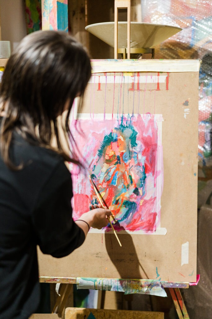 An artist, seen from behind, works on an expressive and colorful oil painting using Pro-Arte brushes. The canvas rests on an easel in a lively studio filled with artistic energy and splashes of creativity.