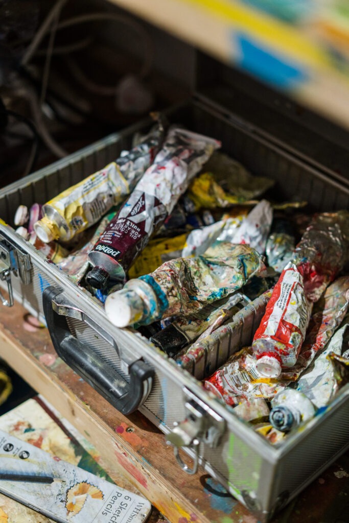 A close-up of a metal box filled with well-used oil paint tubes, showcasing the vibrant colors and messy charm of an artist's tools.