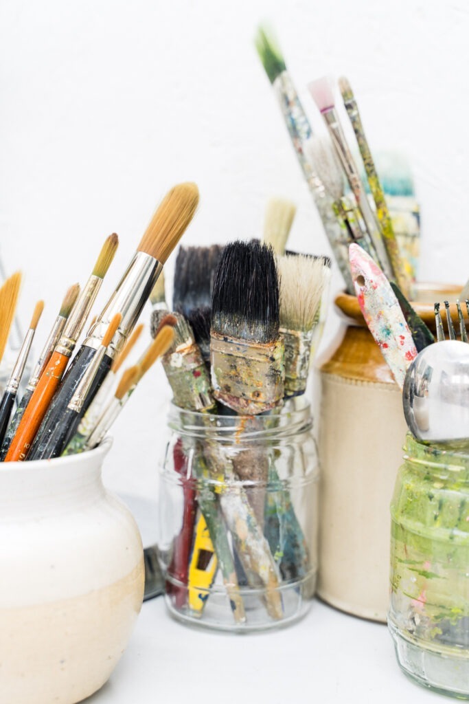 A collection of various paintbrushes and utensils in jars and containers. They are organized standing up with bristles covered in dried paint. The containers are white and ceramic, and the background is a plain white wall.