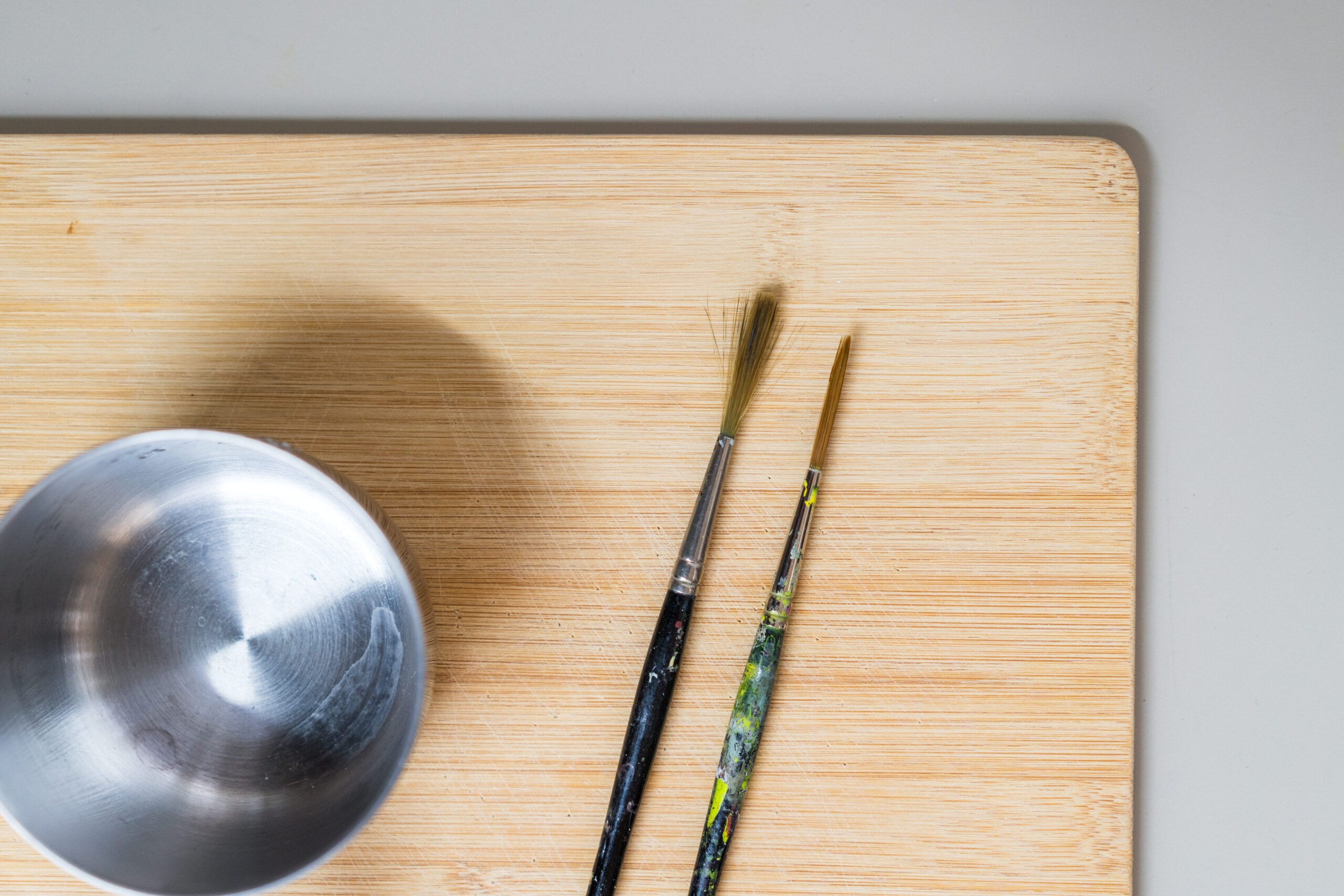 A small metal bowl and two paintbrushes with dried paint rests on a light wooden surface.