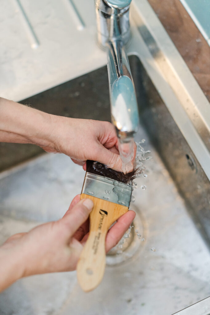 Person washing a Pro Arte paintbrush under a running tap in a stainless steel sink. The brush, with its wooden handle and visible bristles, is being cleaned.