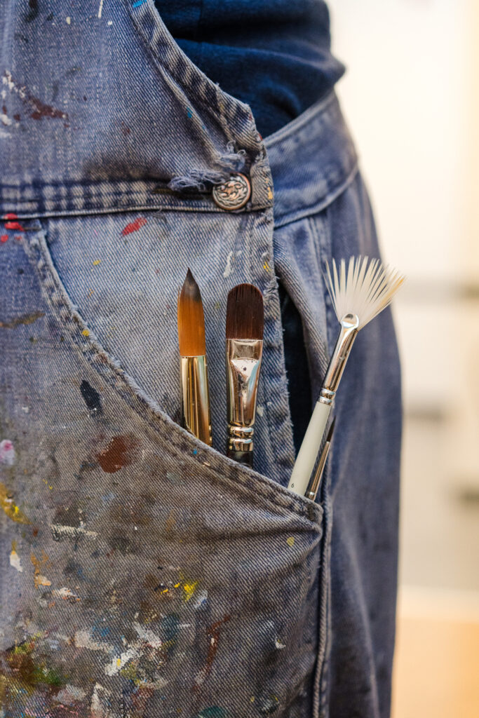 Close-up of a paint-splattered denim apron with three paintbrushes in the pocket.