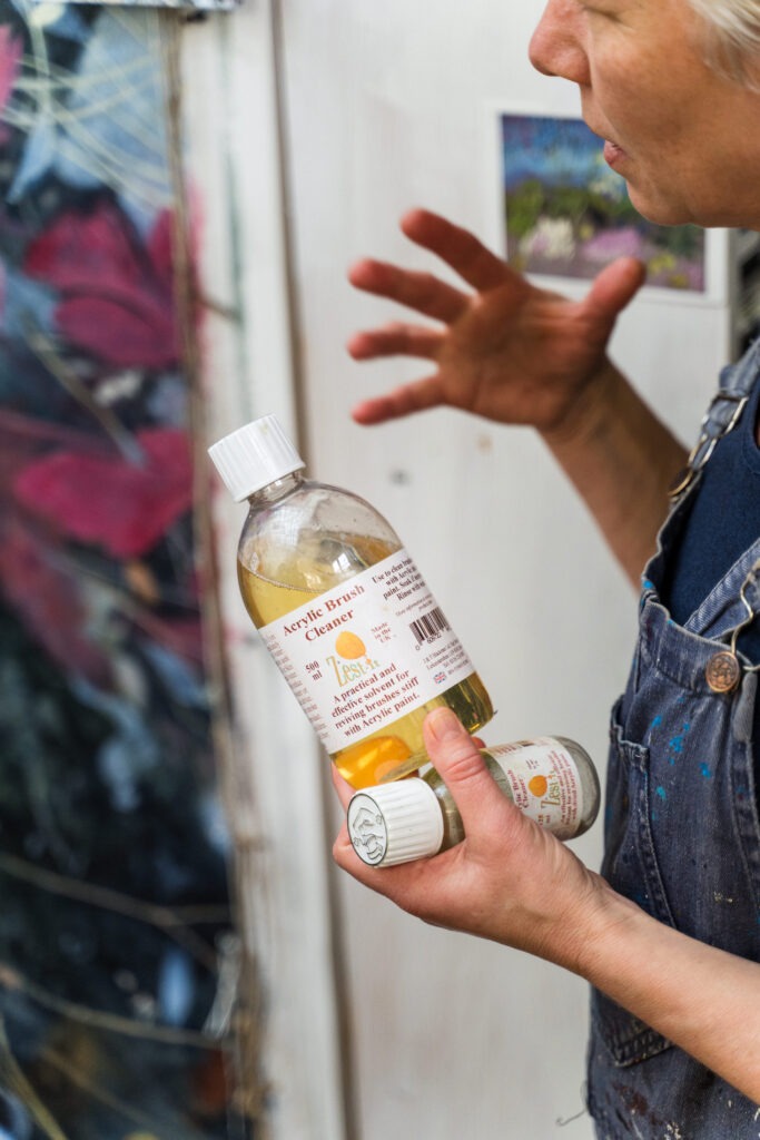 An artist's hands can be seen holding a bottle of cleaning solution for oil paints in a creative studio environment.