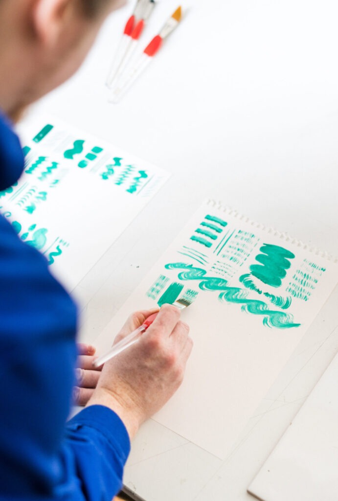 A person wearing a blue shirt is seen from behind, focusing on painting with Pro Arte brushes. They are working with green paint on white paper, creating various brush strokes and patterns. Nearby, several paintbrushes and other painted sheets are visible on the table.