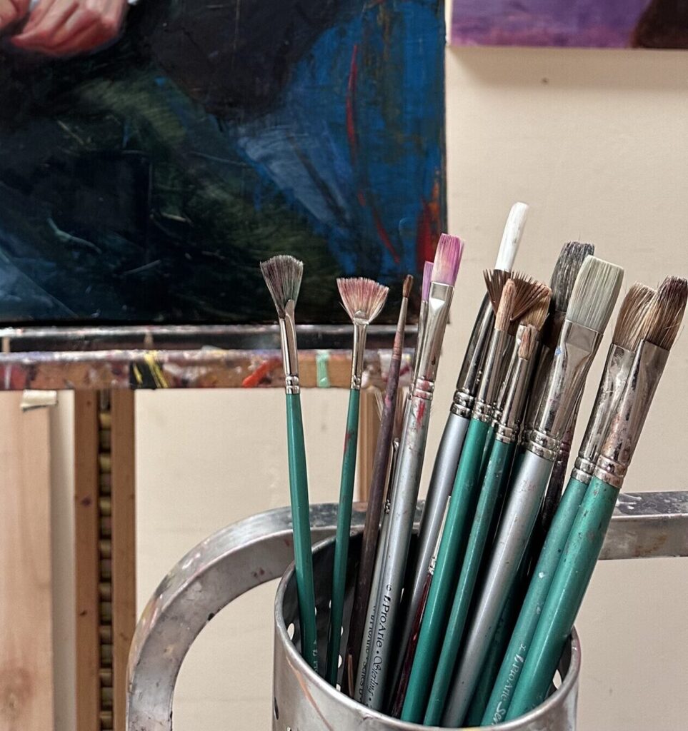 Close-up of a collection of Pro Arte paintbrushes with green and metallic handles, stored in a metal container in an artist's studio. An unfinished painting with dark and vibrant colors is visible in the background on an easel, suggesting an active workspace.