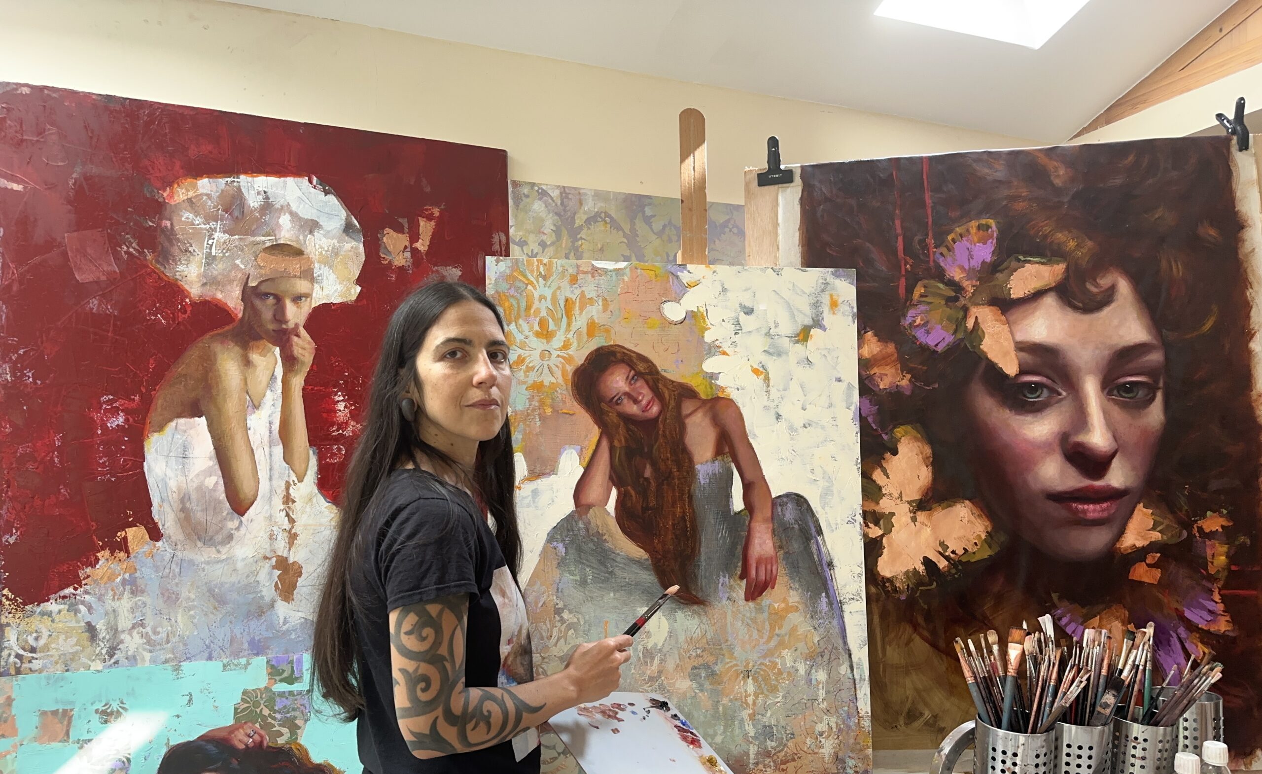 Artist Rosso Crimson standing in her studio, holding a paintbrush and palette while working on a painting of a woman with long hair seated against a textured background. Several of her completed and in-progress paintings, featuring expressive portraits of women, are displayed on easels and walls behind her. The studio is filled with rich colors and dynamic compositions, showcasing her distinctive artistic style.