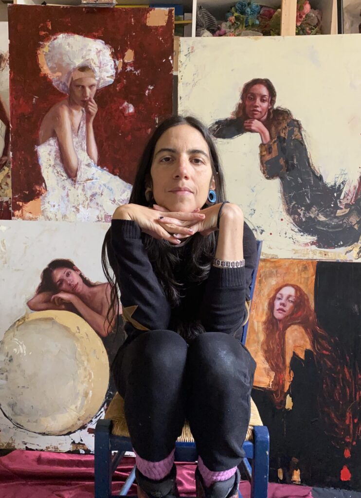 Photograph of an artist seated on a chair with her hands resting under her chin. She is surrounded by her paintings, which depict expressive portraits of women in various poses with rich textures and warm, earthy tones. The vibrant and creative studio space highlights the artist's unique style and dedication to her craft.