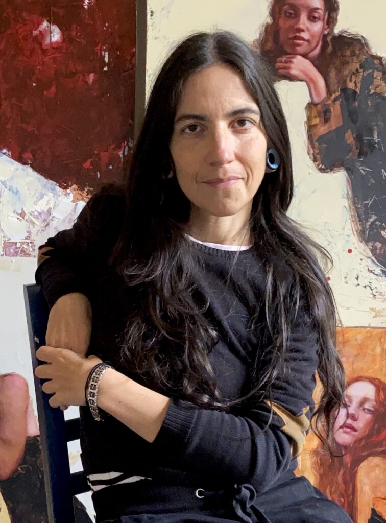 Photograph of an artist seated on a chair with one arm resting on the chair behind her and her hands clasped together. She is surrounded by her paintings, which depict expressive portraits of women in various poses with rich textures and warm, earthy tones. The vibrant and creative studio space highlights the artist's unique style and dedication to her craft.