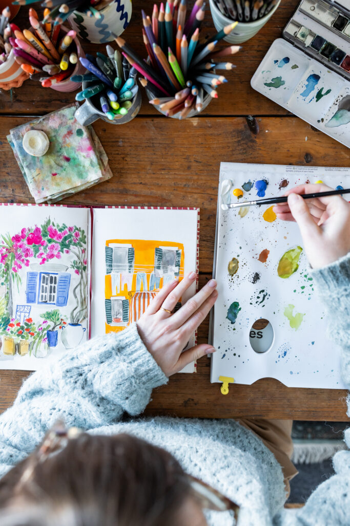 Emma Block, artist and illustrator, painting in a sketchbook with gouache paints. One hand holds the page open to a Mediterranean-style window scene, while the other holds a Pro Arte Artist Paint Brush over a white palette with dabs of paint in various colors.