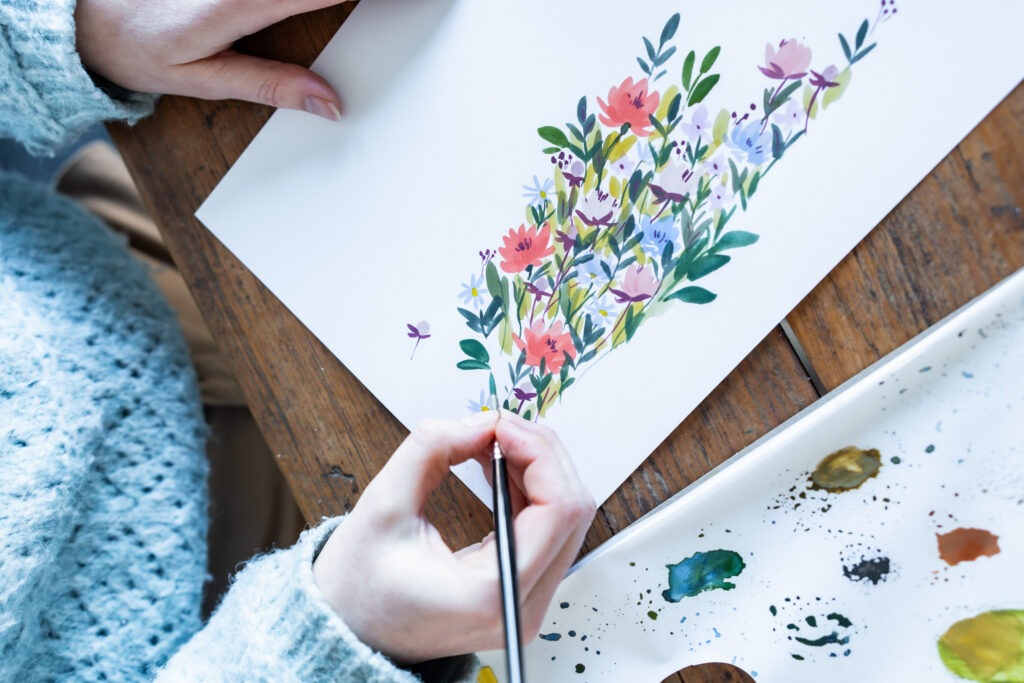 Emma Block painting a bunch of colourful wildflowers in gouache, using a fine Pro Arte Artist Paint Brush. The floral design features red, lilac, blue, and orange blooms with leafy greenery, placed on a wooden desk alongside a paint-splattered palette.