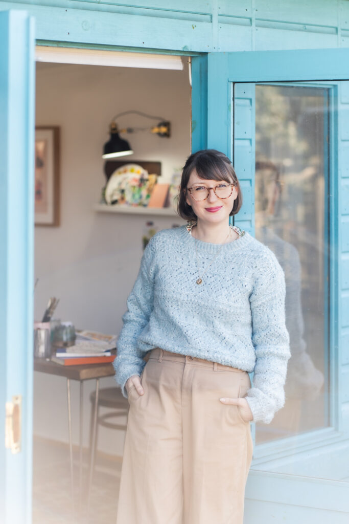 Emma Block, the artist, standing in the doorway of her cozy, sunlit studio. She is smiling and wearing a pale blue knitted jumper and tan trousers, with shelves of her colorful artwork behind her, all painted using her Pro Arte Artist Paint Brushes.