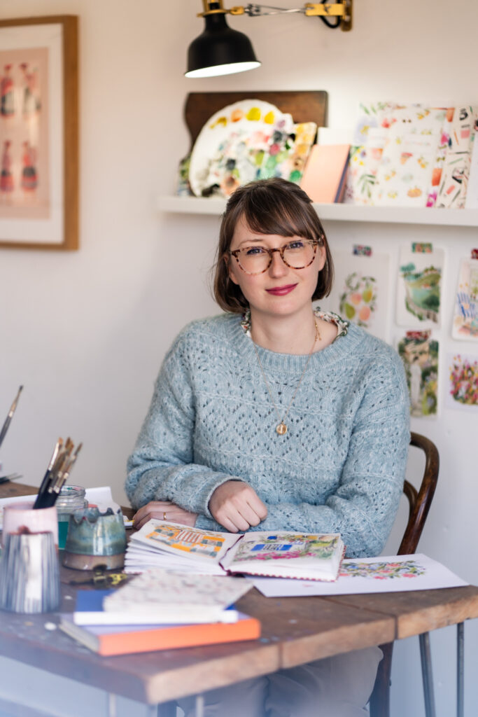 Artist Emma Block seated at her wooden desk, wearing a pale blue textured sweater and glasses. In front of her are her gouache paintings, created with the Pro Arte Emma Block Artist Paint Brushes, and a palette of vibrant paints.