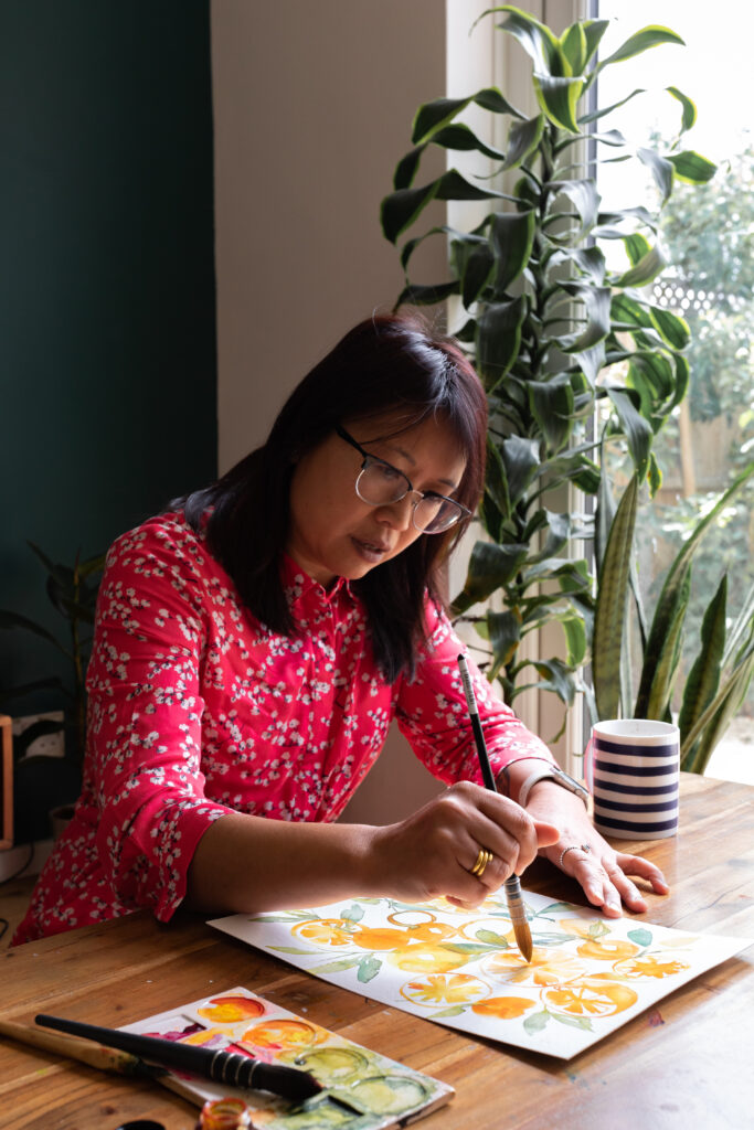 Ohn Mar Win, artist, painting a vibrant orange and green watercolour illustration at a desk surrounded by leafy houseplants. She uses a Pro Arte paint brush, with a striped blue and white mug beside her.