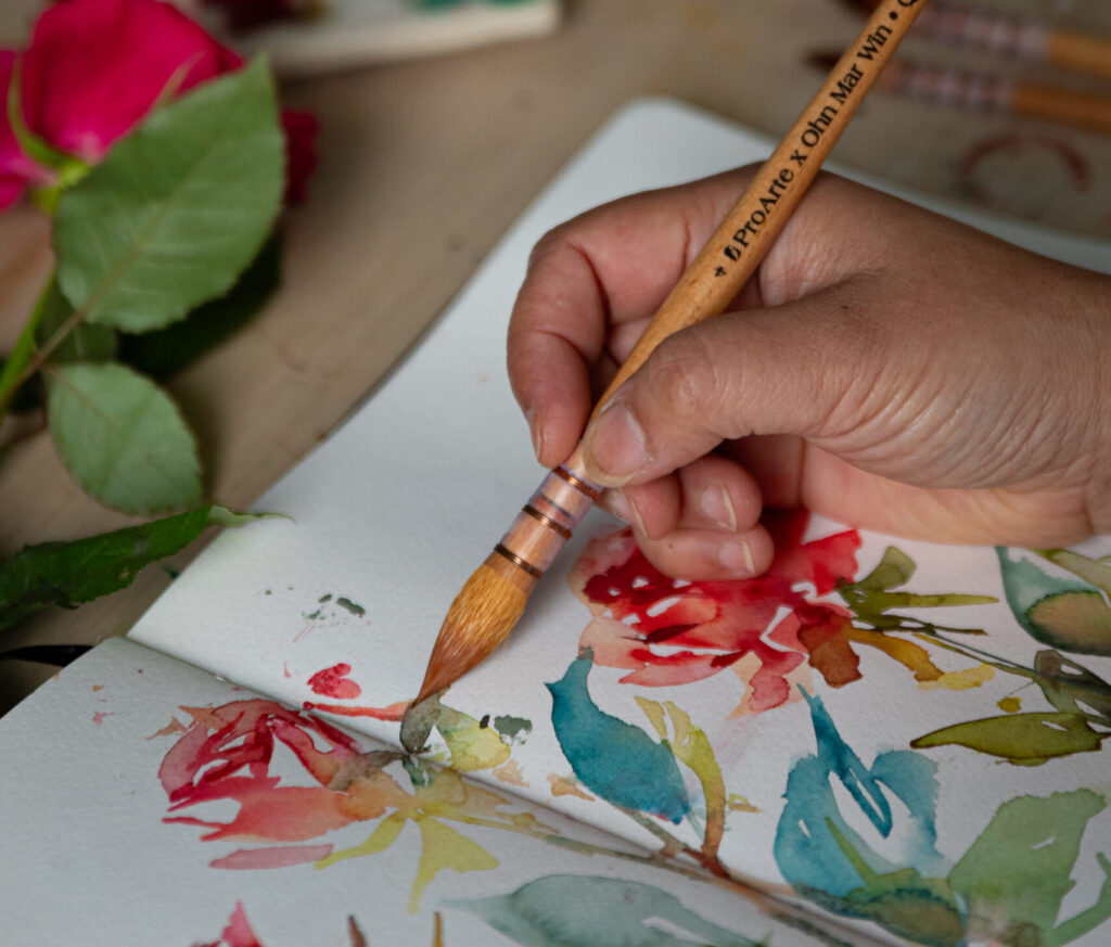 Close-up of artist Ohn Mar Win using a Pro Arte x Ohn Mar Win quill watercolour brush to paint bold, flowing flower shapes in red, teal, and green on textured watercolour paper.