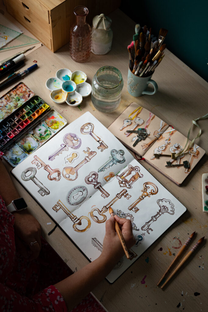 Top-down view of Ohn Mar Win painting a detailed watercolour sketchbook spread filled with antique-style keys. Her Pro Arte paint brushes, a mug of pencils, a glass of water, and watercolour palettes are arranged around her workspace.