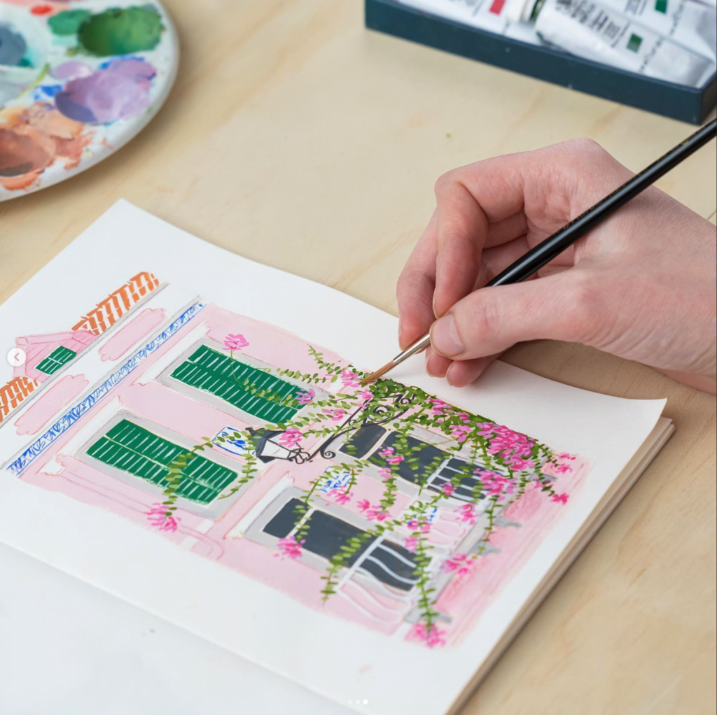 Close-up of artist Emma Block's hand using a fine-tipped Pro Arte Artist Paint Brush to paint delicate green vines and pink flowers on a gouache illustration of a pink house with green shutters.