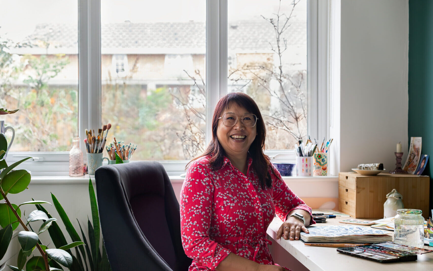 Artist Ohn Mar Win smiling warmly while seated at her bright studio desk, wearing a vibrant red patterned dress. Behind her, large windows reveal a soft, tree-filled view. Her workspace is filled with paint brushes, sketchbooks, and art materials, reflecting her creative process.