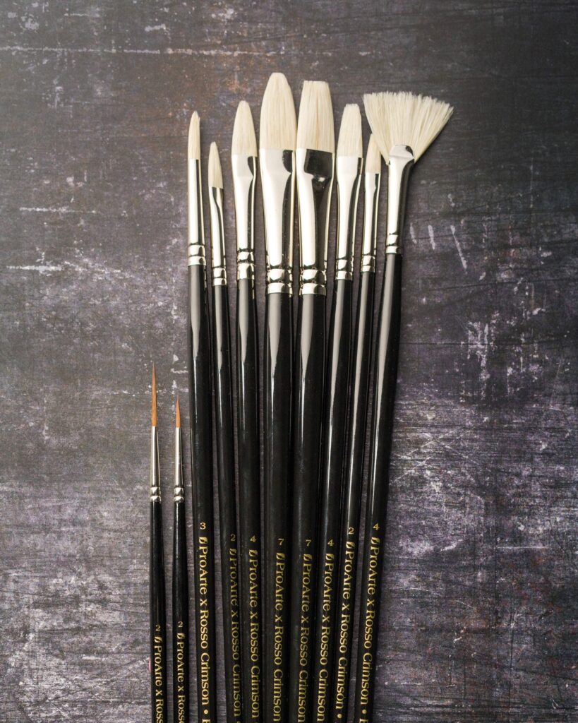 Rosso Crimson x Pro Arte range of hog hair fan, filbert and flat brushes arranged in a row. There are two smaller synthetic brushes placed next to them.